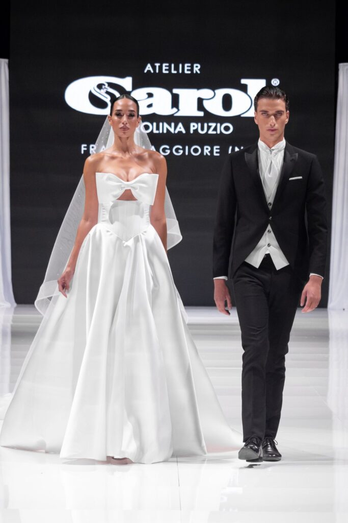 Sposa con abito a fiocco e sposo in tight alla sfilata Atelier Carol