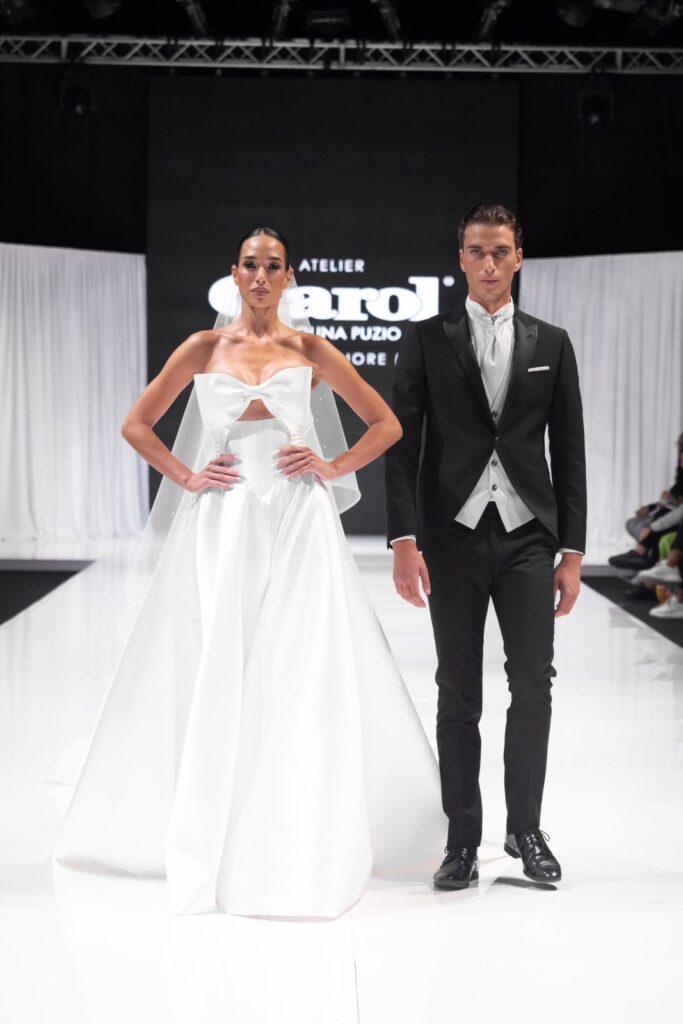 Sposa con fiocco e velo e sposo in tight, sfilata Atelier Carol
